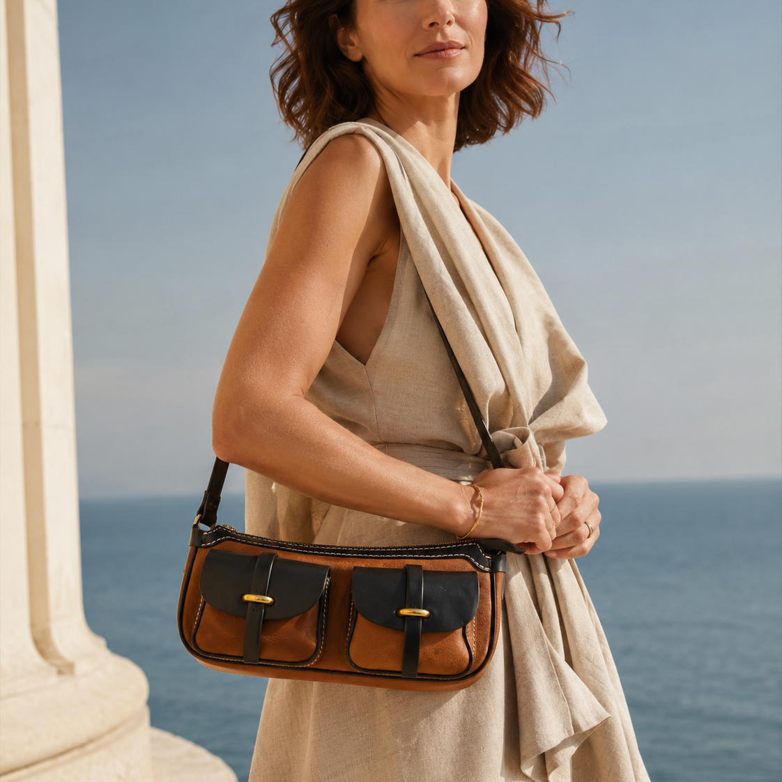Woman wearing a beige sleeveless dress carrying a Dual-Pocket Full-Grain Leather Crossbody Bag in brown and black with vintage brass hardware by the ocean.