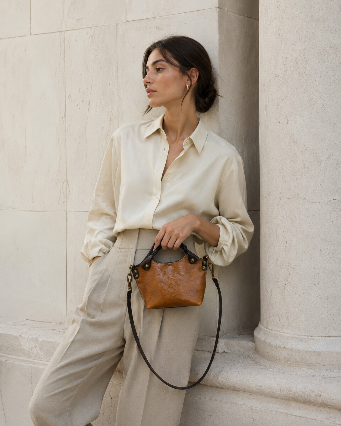 Woman holding a brown leather handbag against a light stone wall.