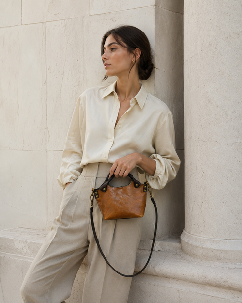 Woman holding a brown leather handbag against a light stone wall.