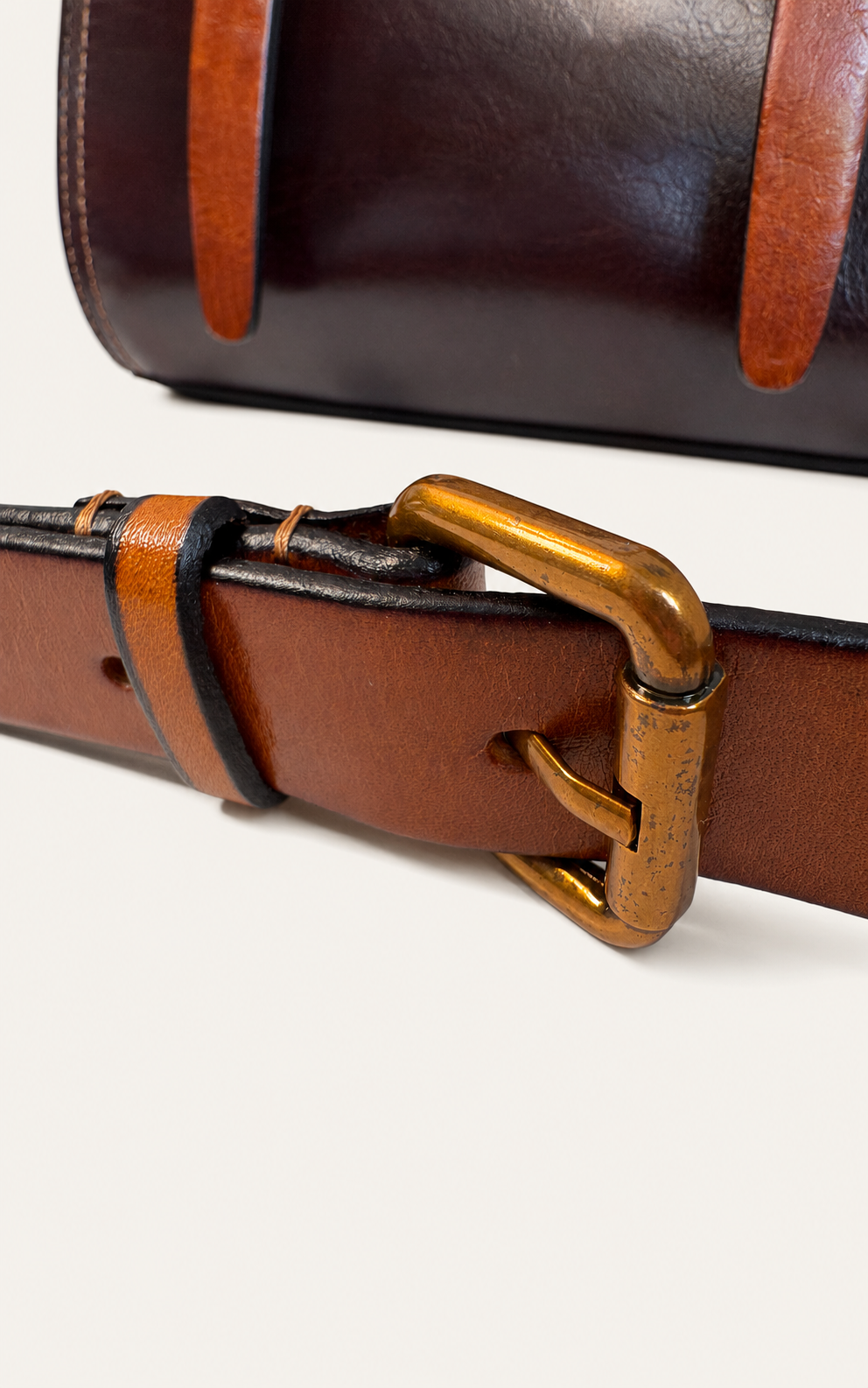 A close-up of a brown leather belt with a gold-tone buckle, shown against a light neutral background with a second dark brown leather item partially visible at the top.