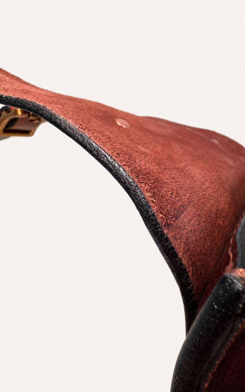 Close-up of a reddish-brown leather strap or saddle strap with black edging and a small brass buckle, shown against a plain light background.