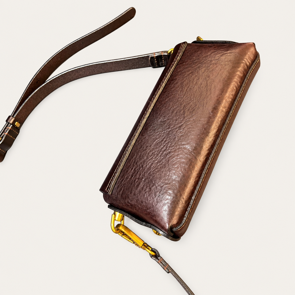 A brown leather wristlet clutch with a matching leather strap and gold-tone hardware, shown on a light off-white background.