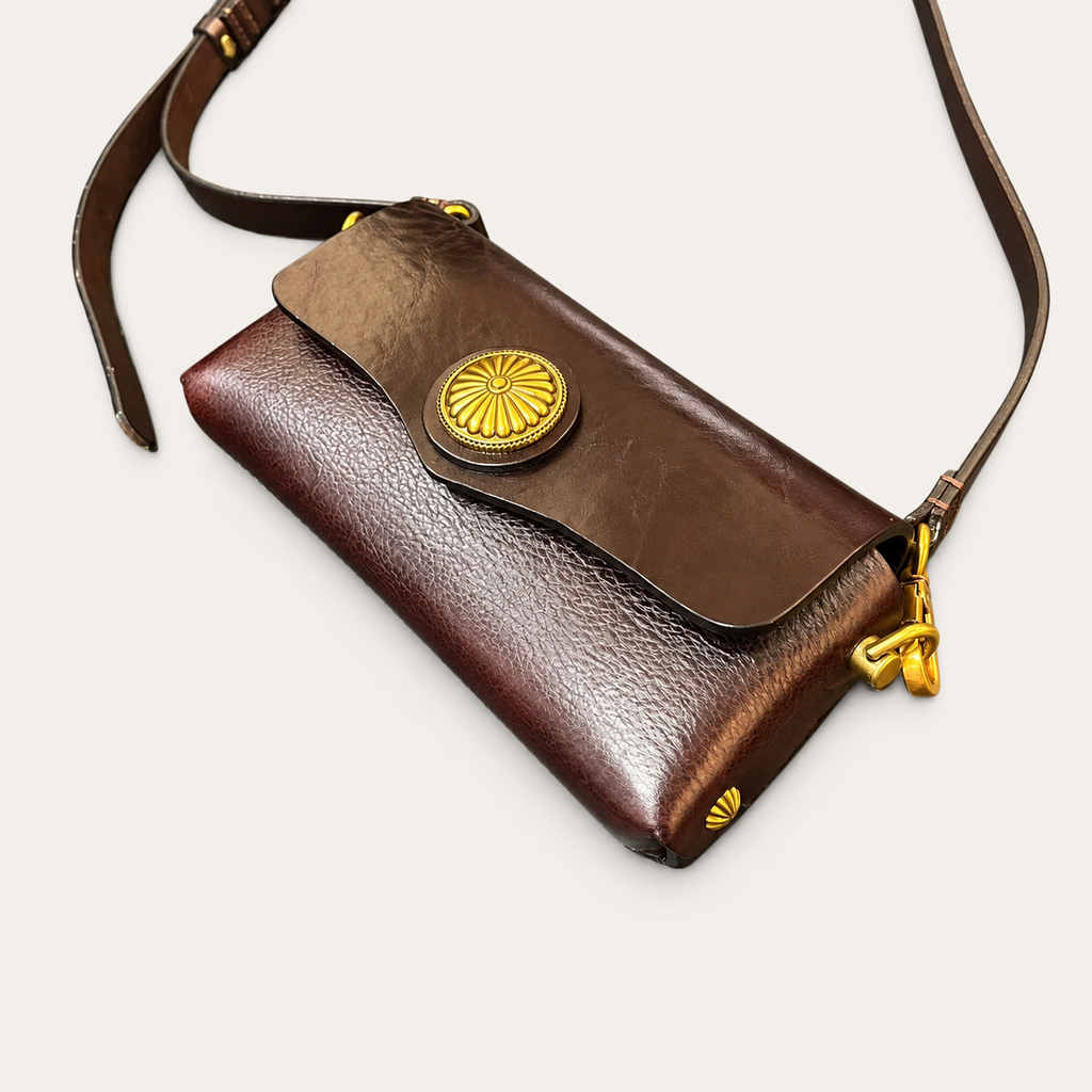A dark brown leather crossbody bag with a textured finish, a gold-toned circular flower-shaped clasp, and a matching gold-toned keyring detail on the strap, shown on a light off-white background.