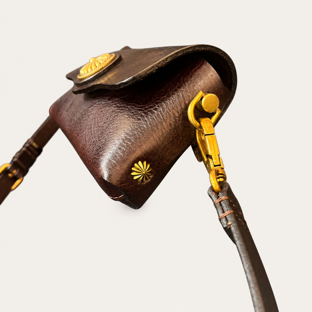A close-up of a dark brown leather shoulder bag with gold-tone hardware, featuring a textured leather flap with a decorative gold emblem and a matching gold clasp connected to a dark brown strap, set against a solid off-white background.