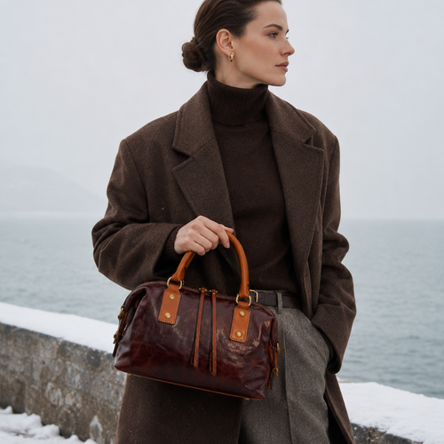 Woman in a brown coat holding a brown leather bag by a snowy waterfront.