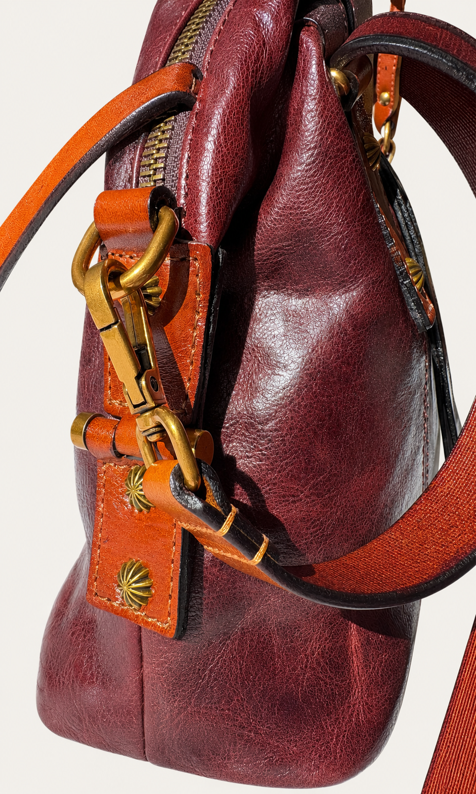 Close-up of a burgundy leather handbag with orange-brown leather straps and gold-tone hardware on a light beige background.