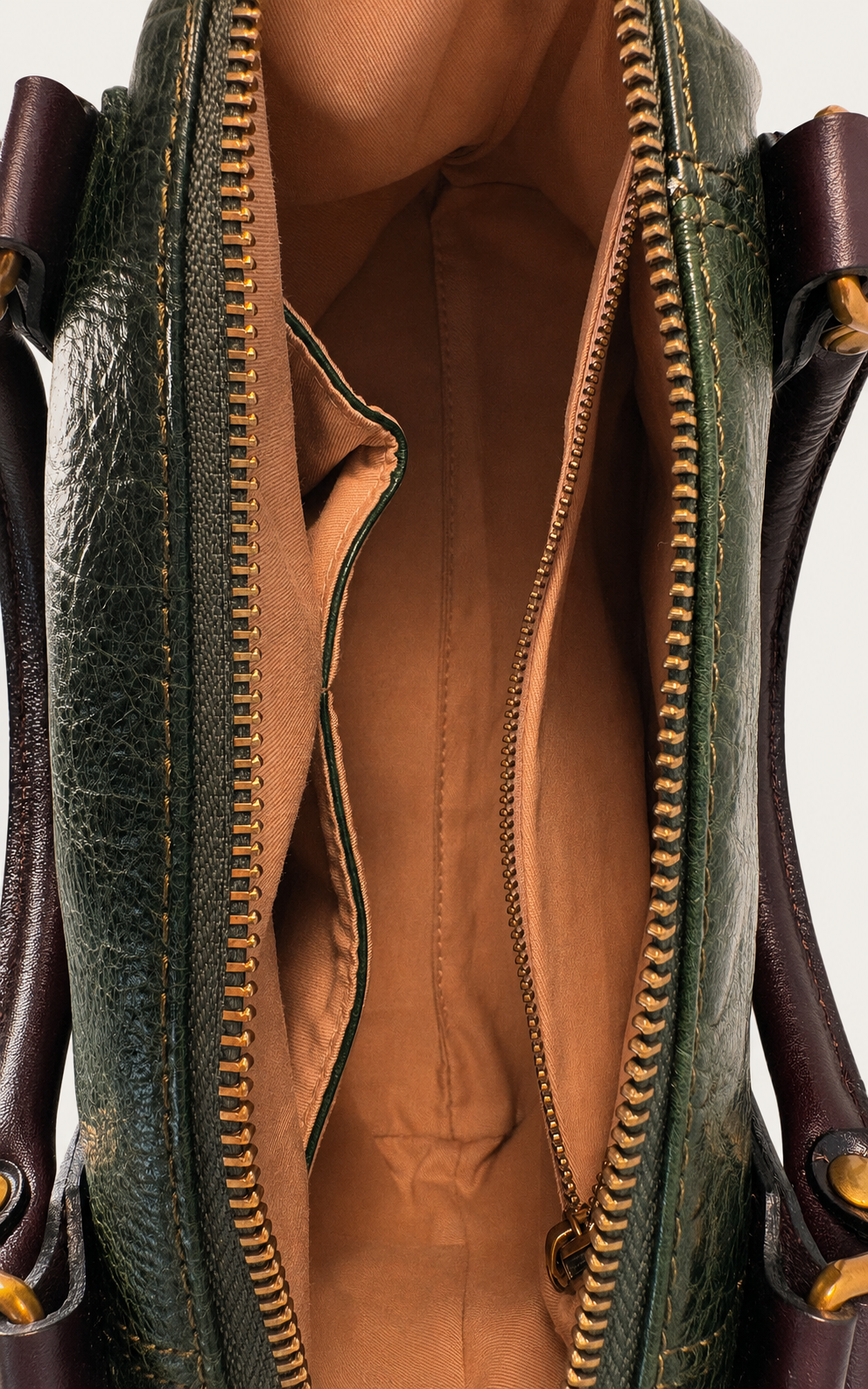 Close-up of an open dark green leather handbag showing a tan interior lining with gold-toned zipper teeth and side handles.