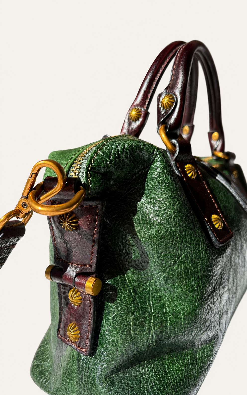Close-up of a green textured leather handbag with dark brown leather handles and straps, accented by gold-tone hardware and decorative starburst stitching.