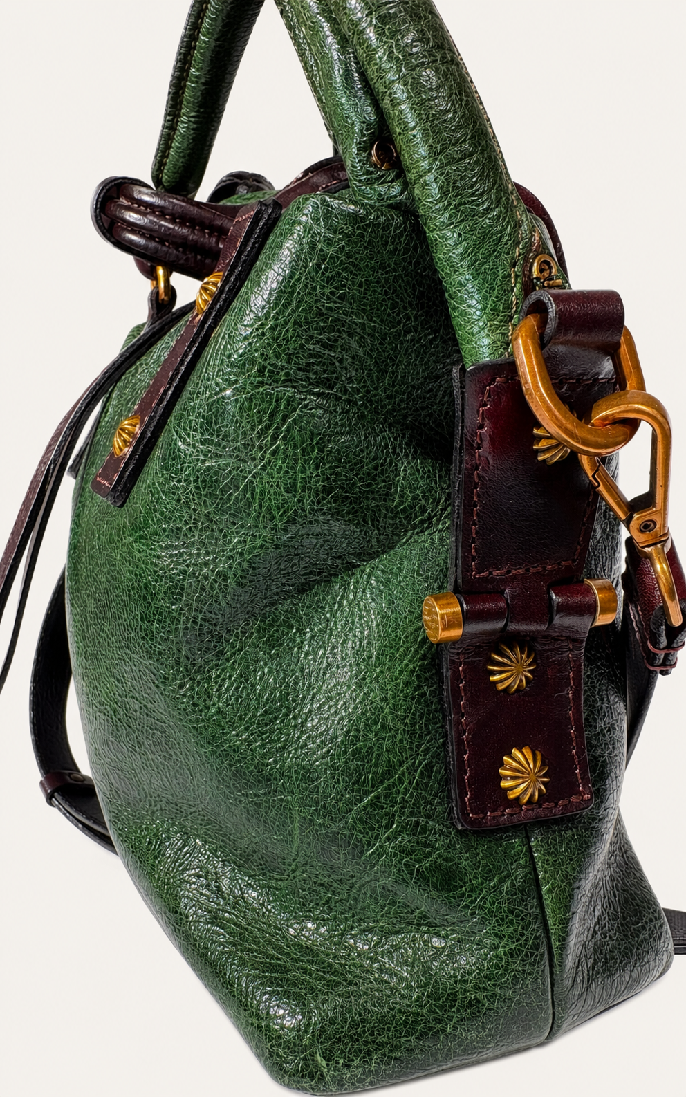 A close-up of a green textured leather handbag with dark brown leather trim and gold-tone hardware, including a prominent ring clasp and decorative starburst studs.