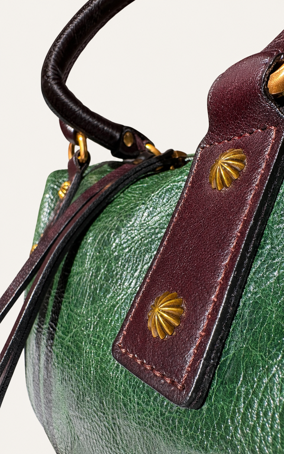 Close-up of a green pebbled leather handbag with dark brown leather trim and gold shell-shaped hardware, showing part of the handle and strap.