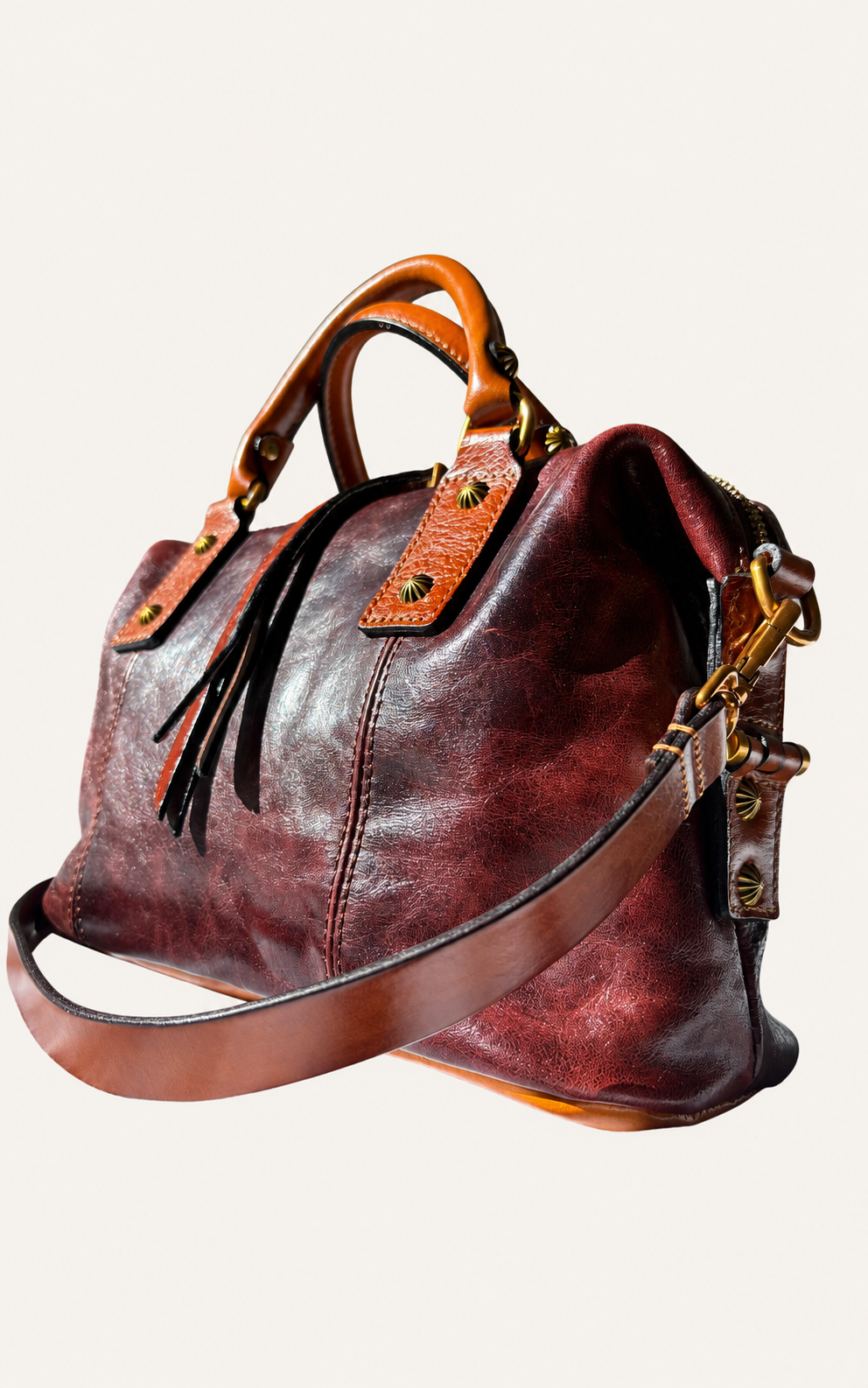 A distressed leather handbag in a deep burgundy red with tan leather handles and strap, accented by gold-tone hardware, shown on a light beige background.