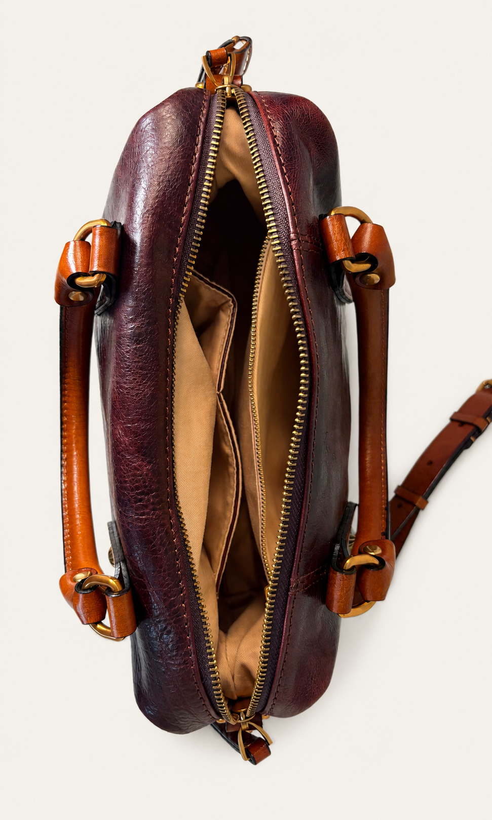 A top-down view of a dark burgundy leather bag with a gold zipper, tan interior, and brown leather handles on a light beige background.