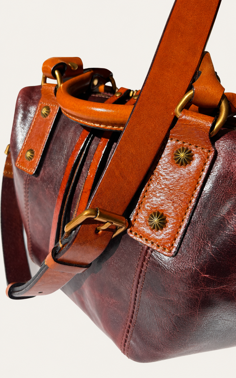 Close-up of a leather handbag in deep burgundy and tan-brown tones with brass-tone hardware and a wide brown shoulder strap against a light neutral background.