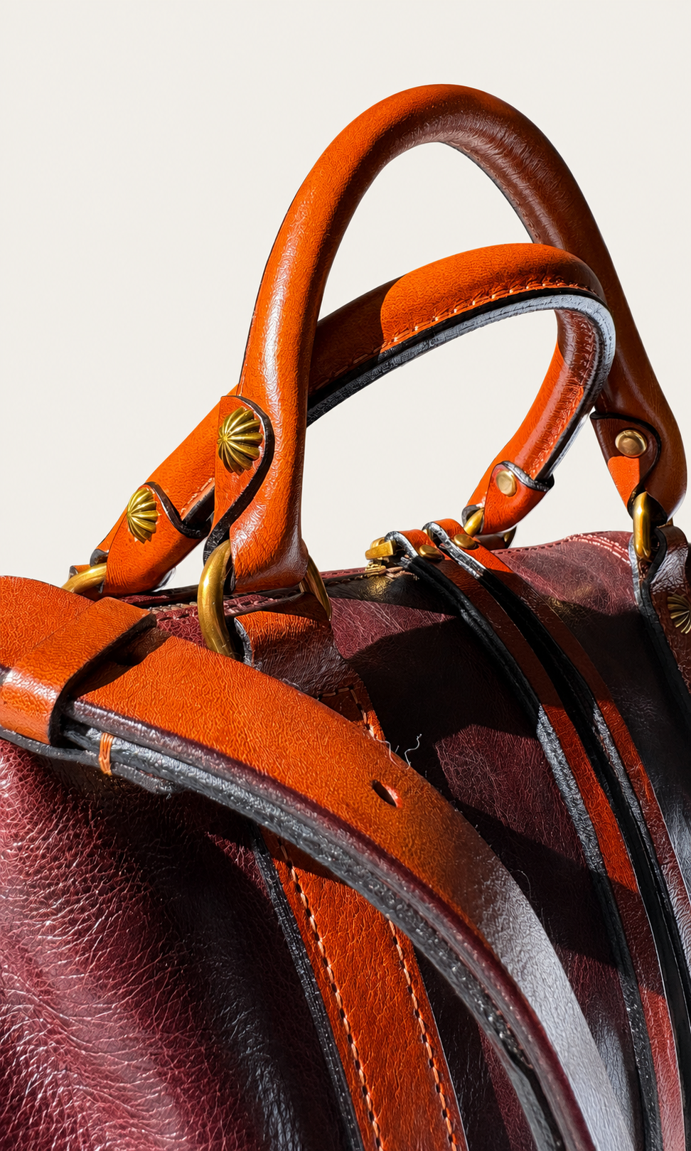 Close-up of a leather handbag showing warm cognac brown handles, a burgundy body, and brass-tone hardware against a light neutral background.