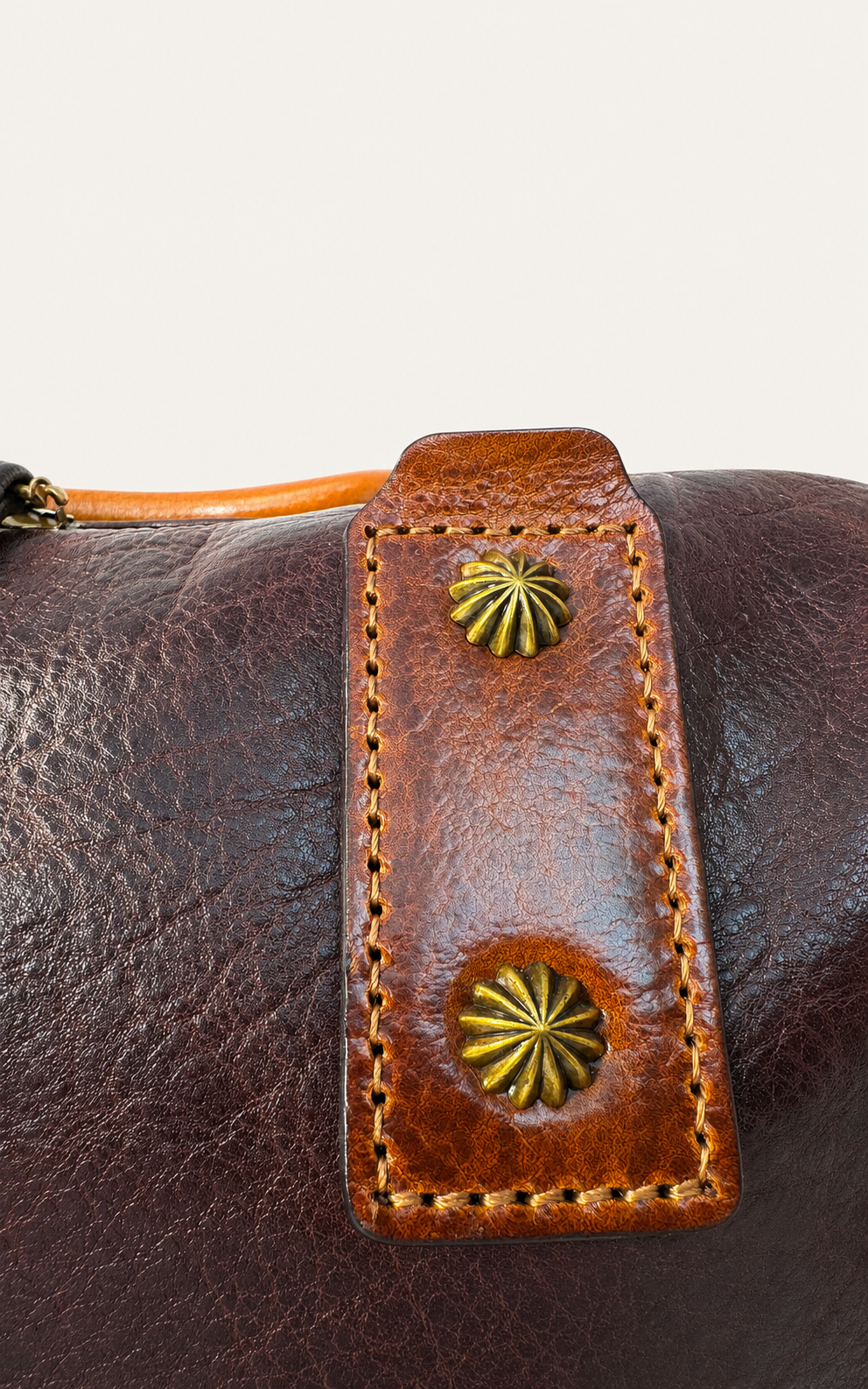 Close-up of a brown leather bag featuring a stitched leather strap closure with two gold-toned starburst studs, set against a plain light background.