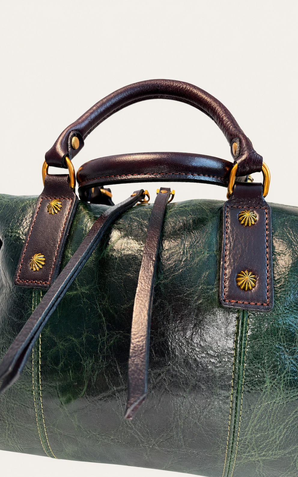 Close-up of a dark green leather duffel bag with dark brown leather handles and trim, accented by gold-tone hardware and decorative starburst studs.