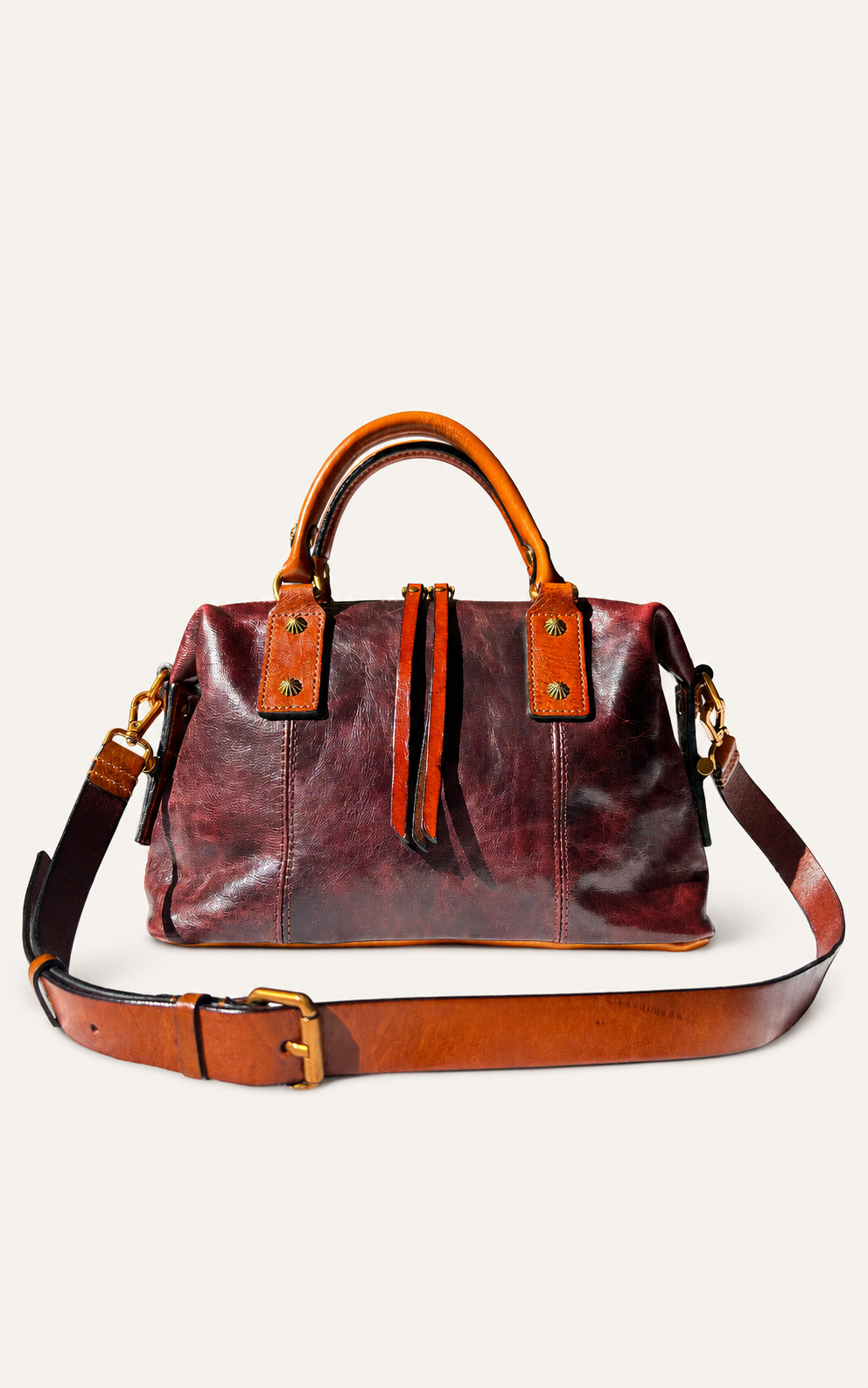 A distressed leather duffel bag in deep burgundy and brown, featuring tan leather top handles, brass-tone hardware, and a matching adjustable shoulder strap laid out in front on a light beige background.