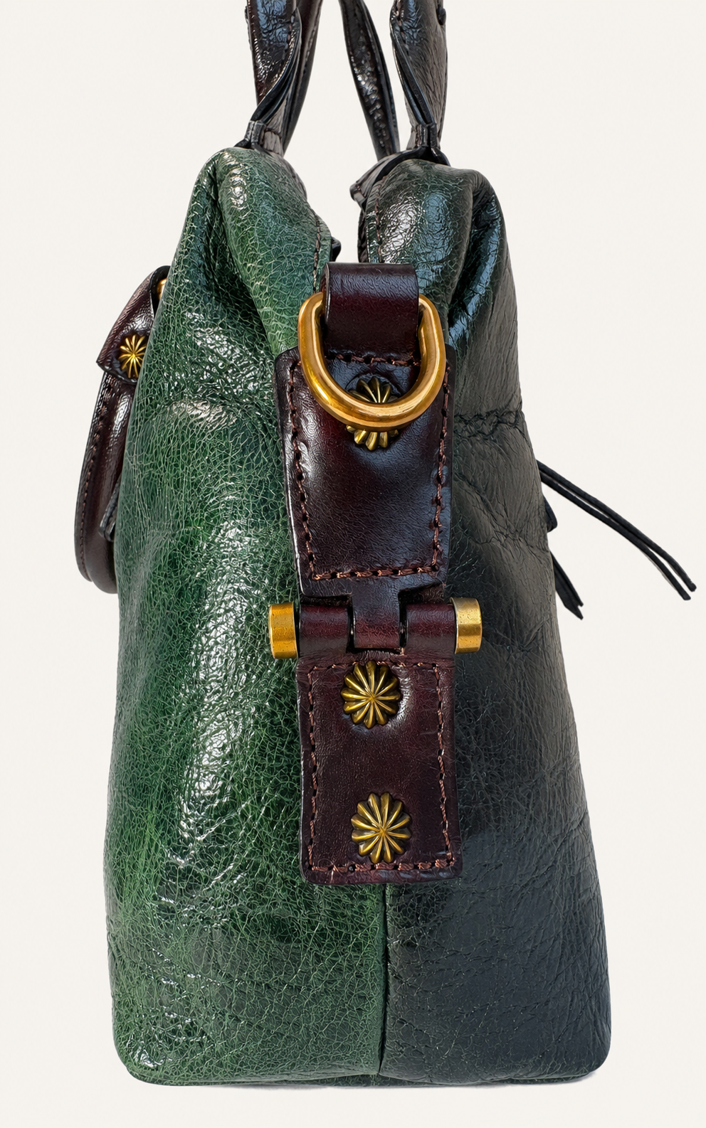 A close-up of a textured leather handbag in green and dark brown, featuring a brown leather strap closure with gold-tone hardware and starburst details, set against a plain off-white background.