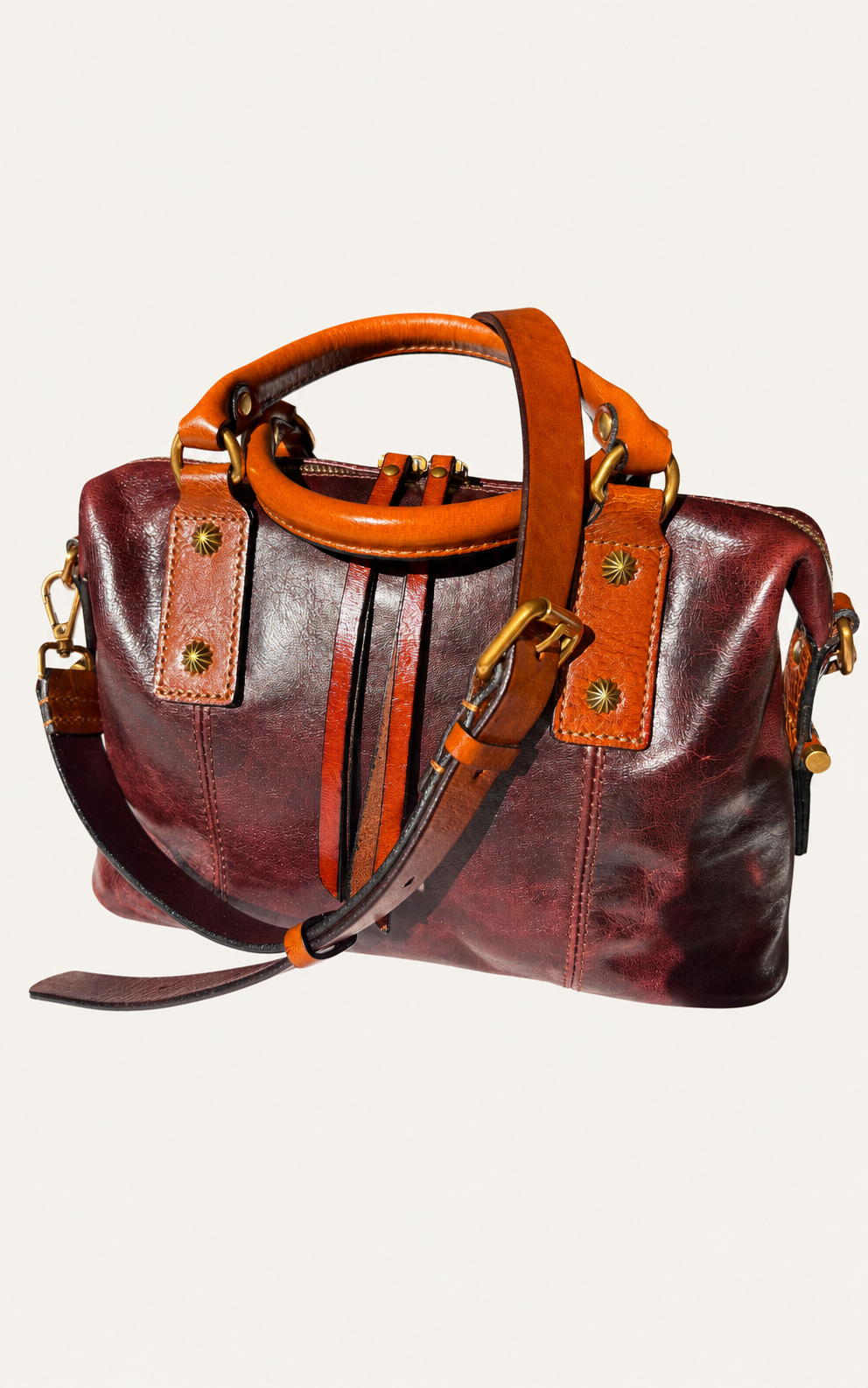 A distressed leather duffle bag in deep burgundy and brown with tan leather handles and straps, and gold-tone hardware, shown against a light beige background.