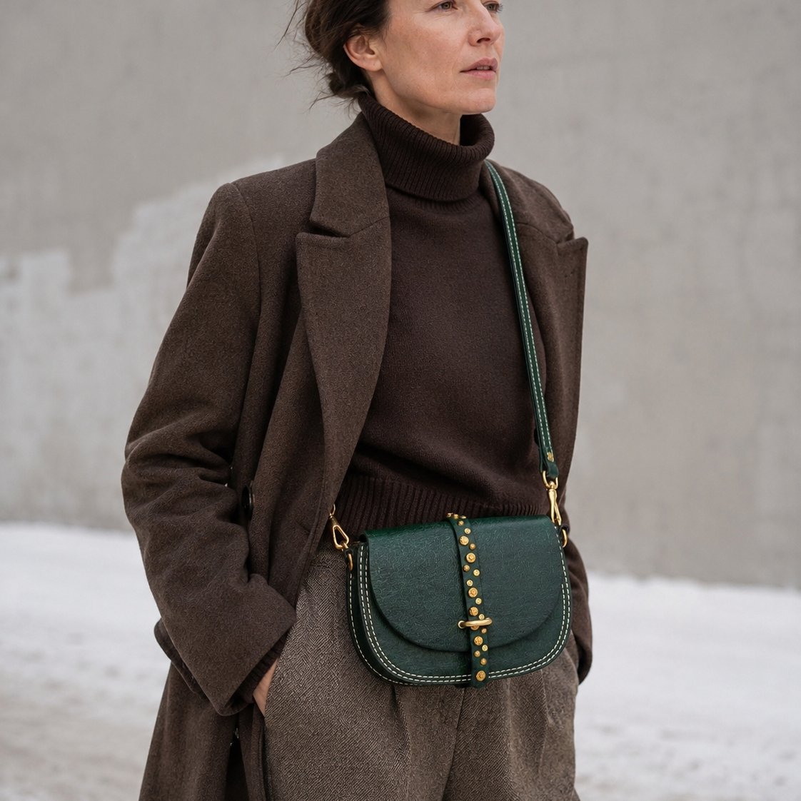 Woman wearing a brown coat and sweater with a green handbag against a neutral background
